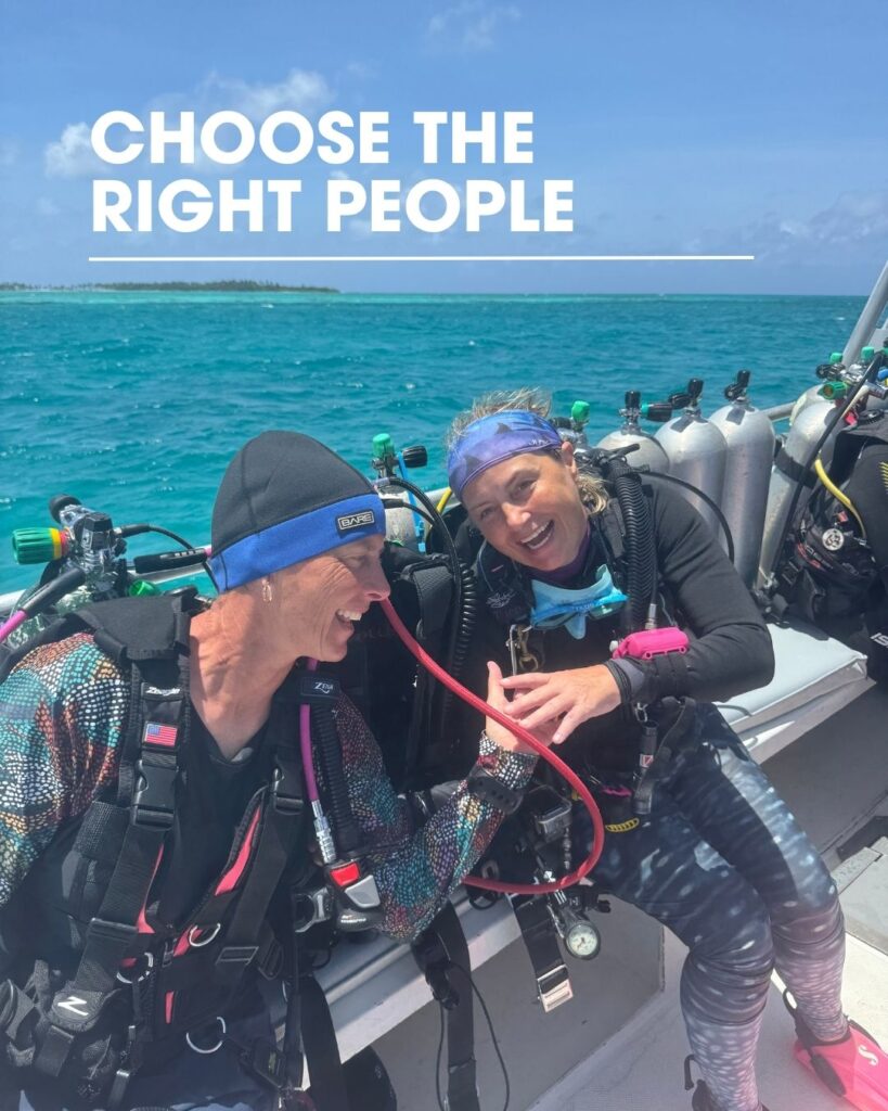Two women scuba divers in full gear laughing together on a dive boat, showing supportive dive buddies
