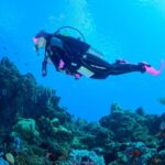 An image of a woman in her fifties in full dive gear diving amidst a coral reef, looking out at the underwater world, composed and present