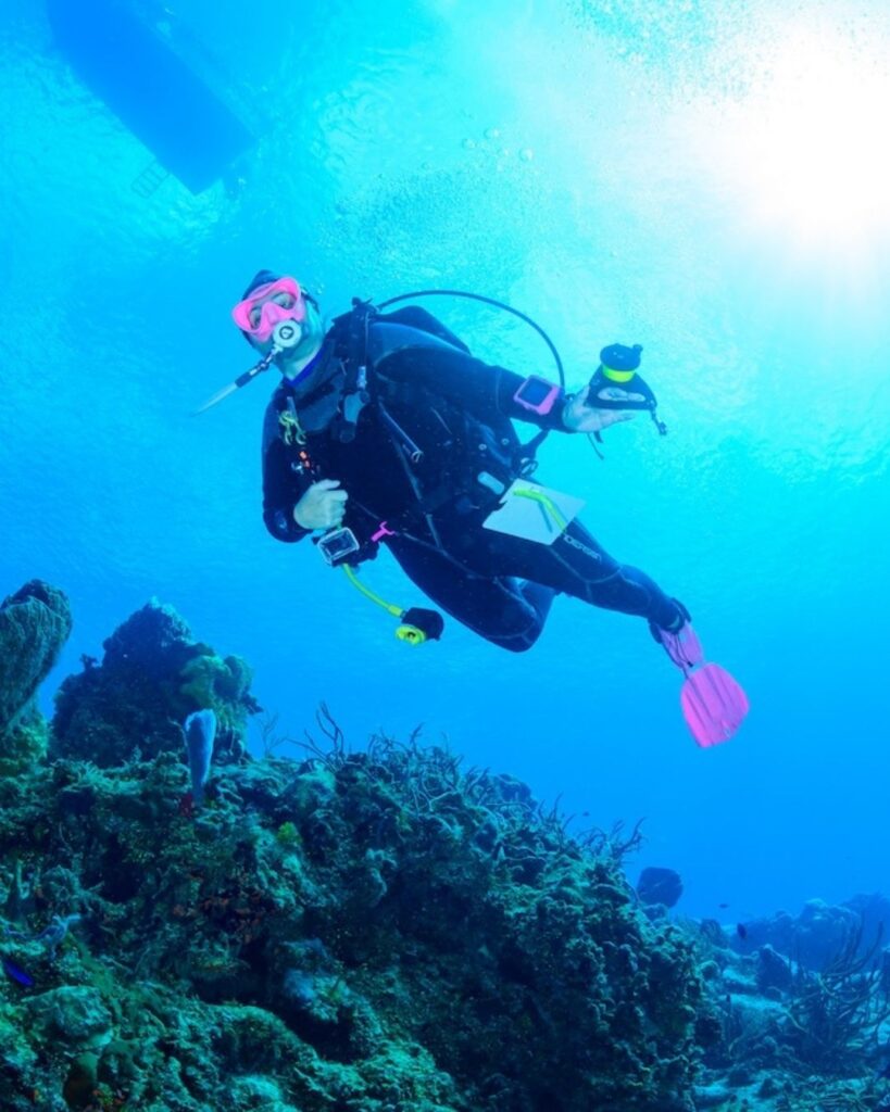 A woman diver hovering mid-water column in neutral buoyancy, light filtering down from the surface, body relaxed and still before reaching the reef
